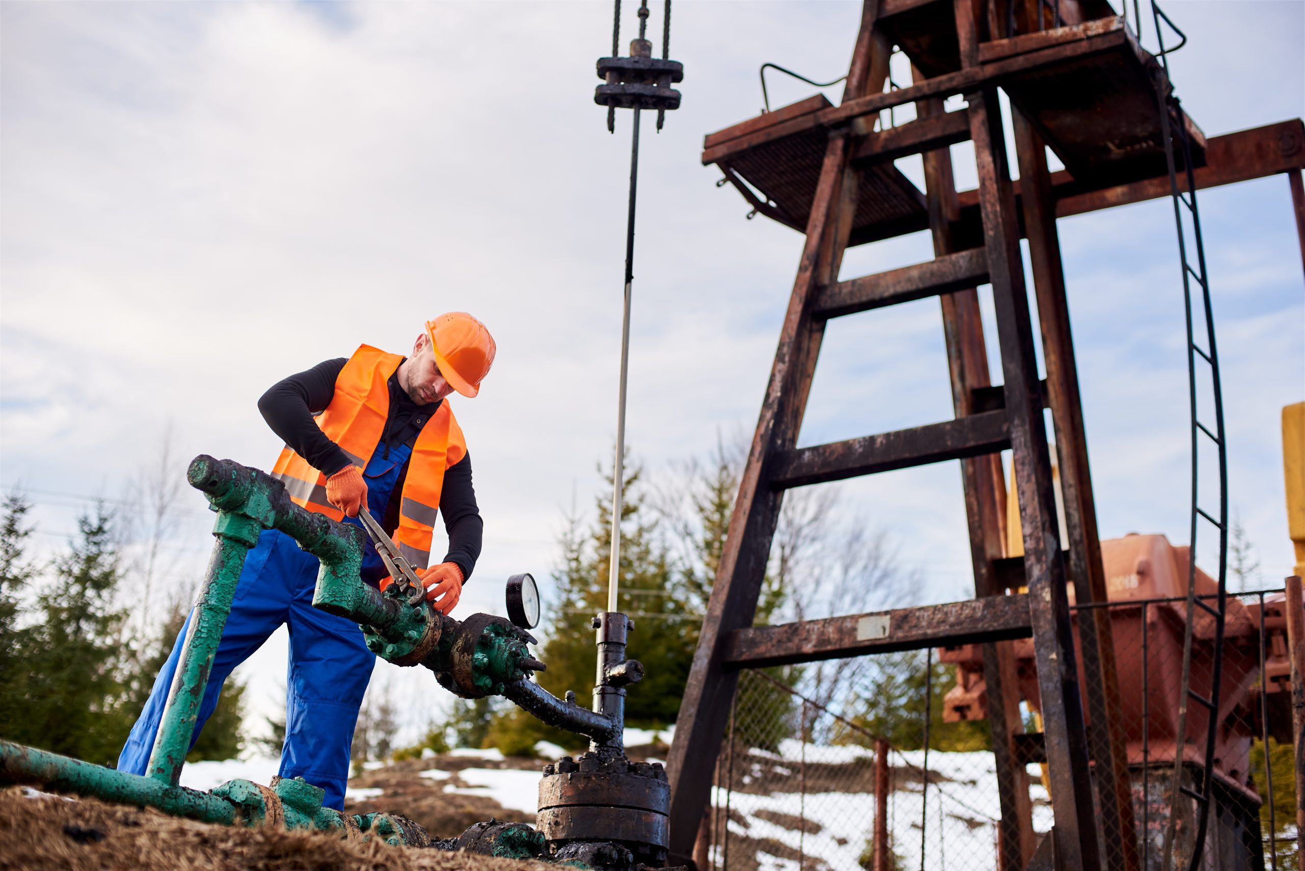 Oil worker in blue overalls, orange vest and helmet working with wrenching a bolt on a gas pipe near an oil pump jack, concept of oil industry